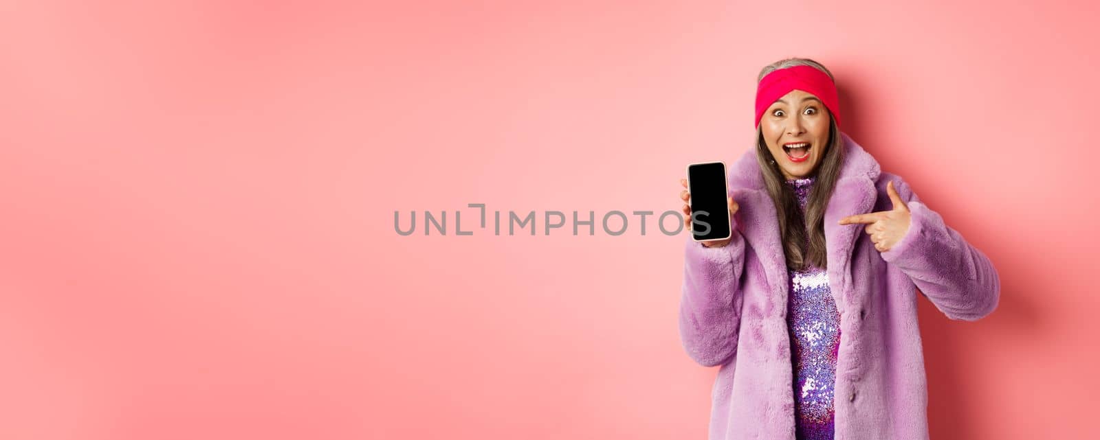 Online shopping and fashion concept. Stylish asian senior woman in faux fur coat pointing finger at blank smartphone screen, smiling happy, showing application on phone by Benzoix