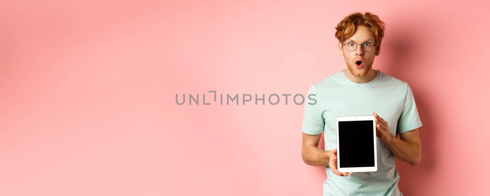 Excited young man with red hair and beard, checking out online promotion, showing digital tablet screen and staring at camera amazed, standing over pink background.