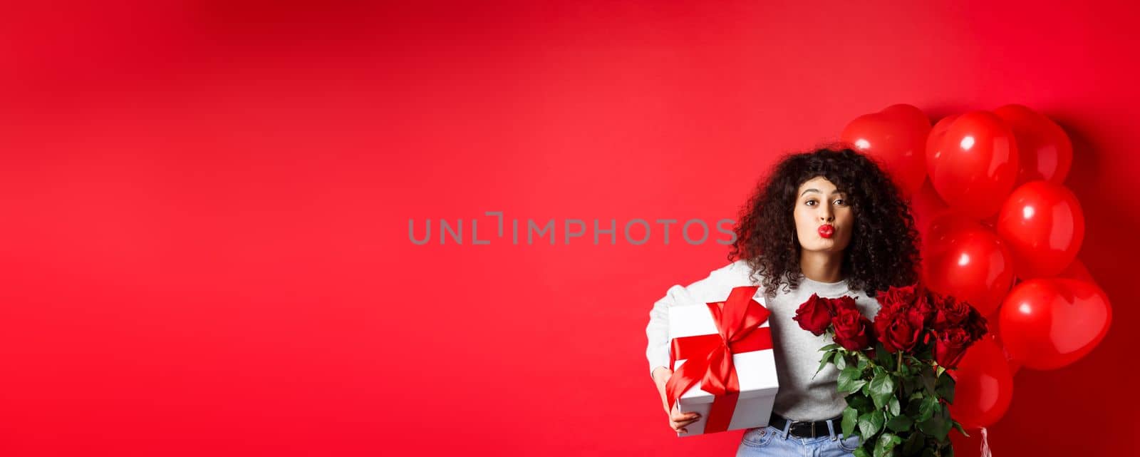 Holidays and celebration. Pretty woman celebrating birthday blowing air kiss, receive gifts and flowers on anniversary, standing near party balloons, red background.