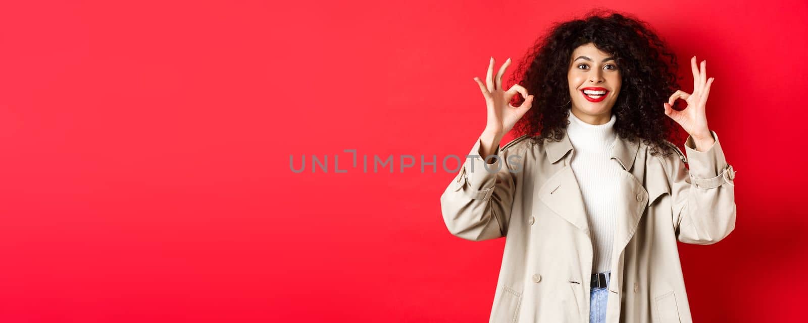 Stylish caucasian woman in trench coat showing okay gesture and smiling satisfied, recommending company, standing on red background.