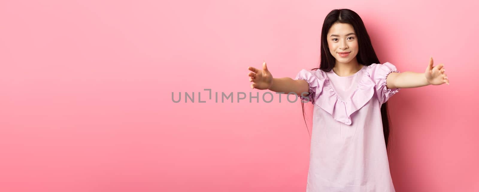 Cute teen asian girl stretch out hands for hug, want to cuddle, friendly greeting you, standing on pink background.