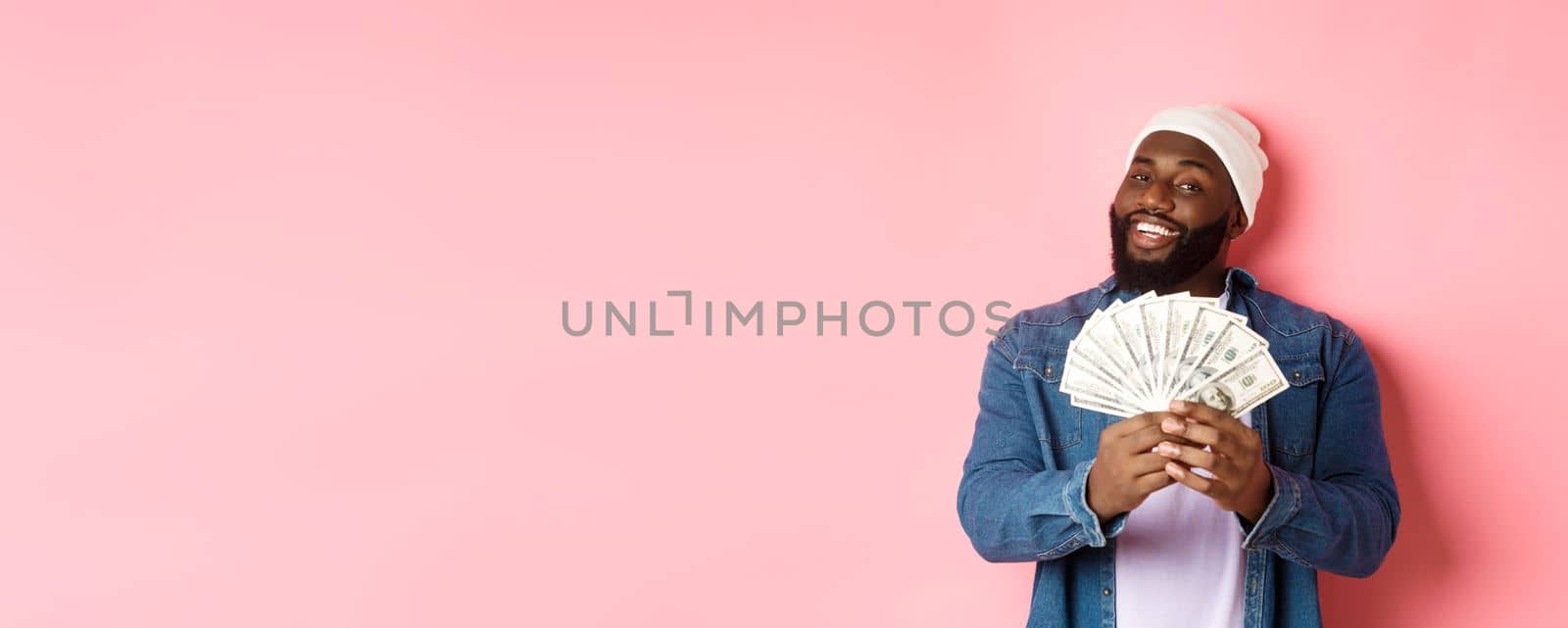 Satisfied rich african-american man showing money, smiling pleased and show-off with income, standing with dollars over pink background by Benzoix