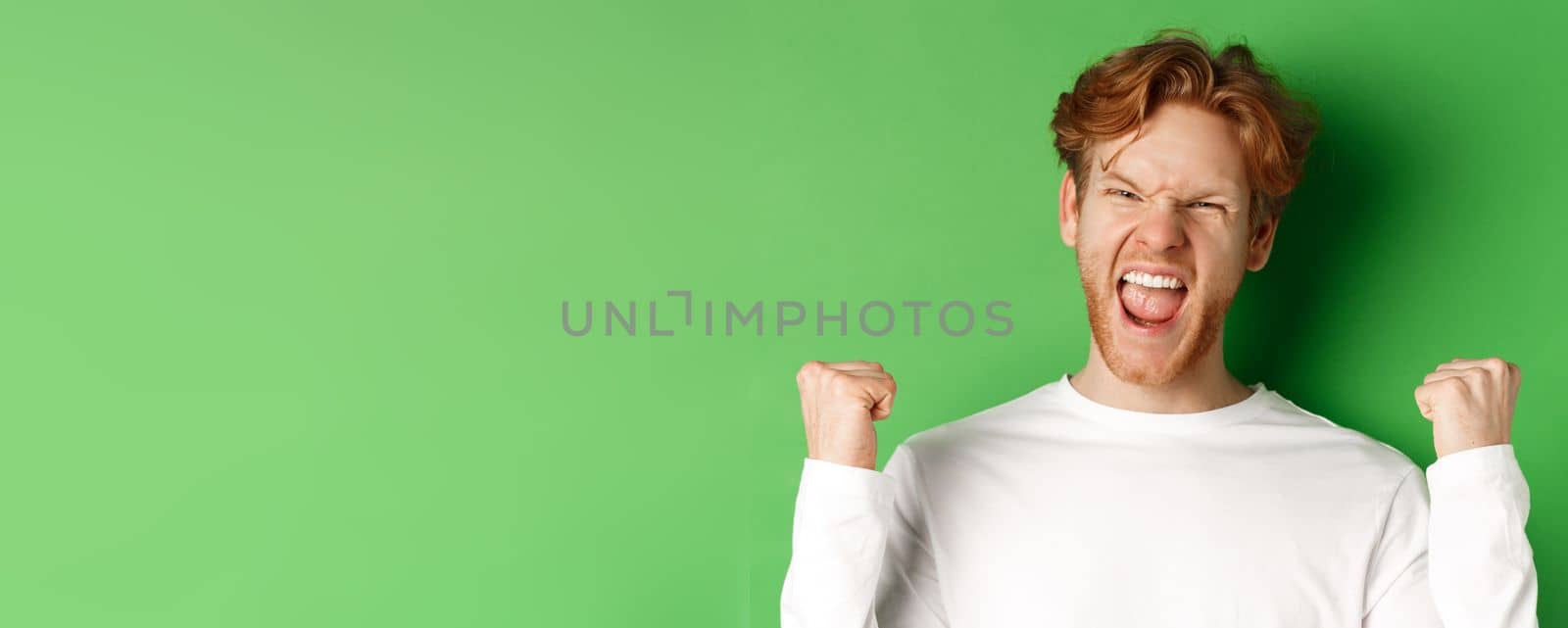 Close-up of lucky young man winner, celebrating victory, shouting yes with joy and making fist pumps, triumphing on green background by Benzoix