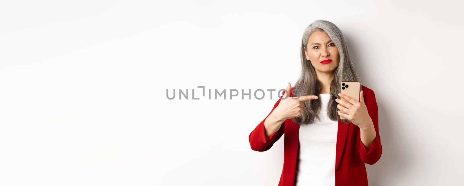 Disappointed mature asian woman complaining, pointing finger at smartphone and looking displeased, standing over white background by Benzoix