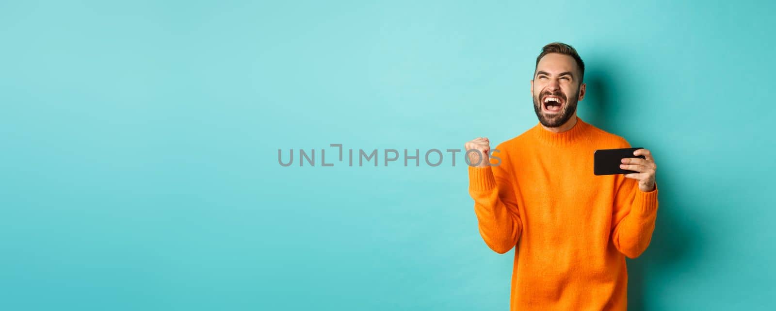 Image of man winning on mobile phone, making fist pump and rejoicing of victory, holding smartphone with satisfied face, turquoise background by Benzoix