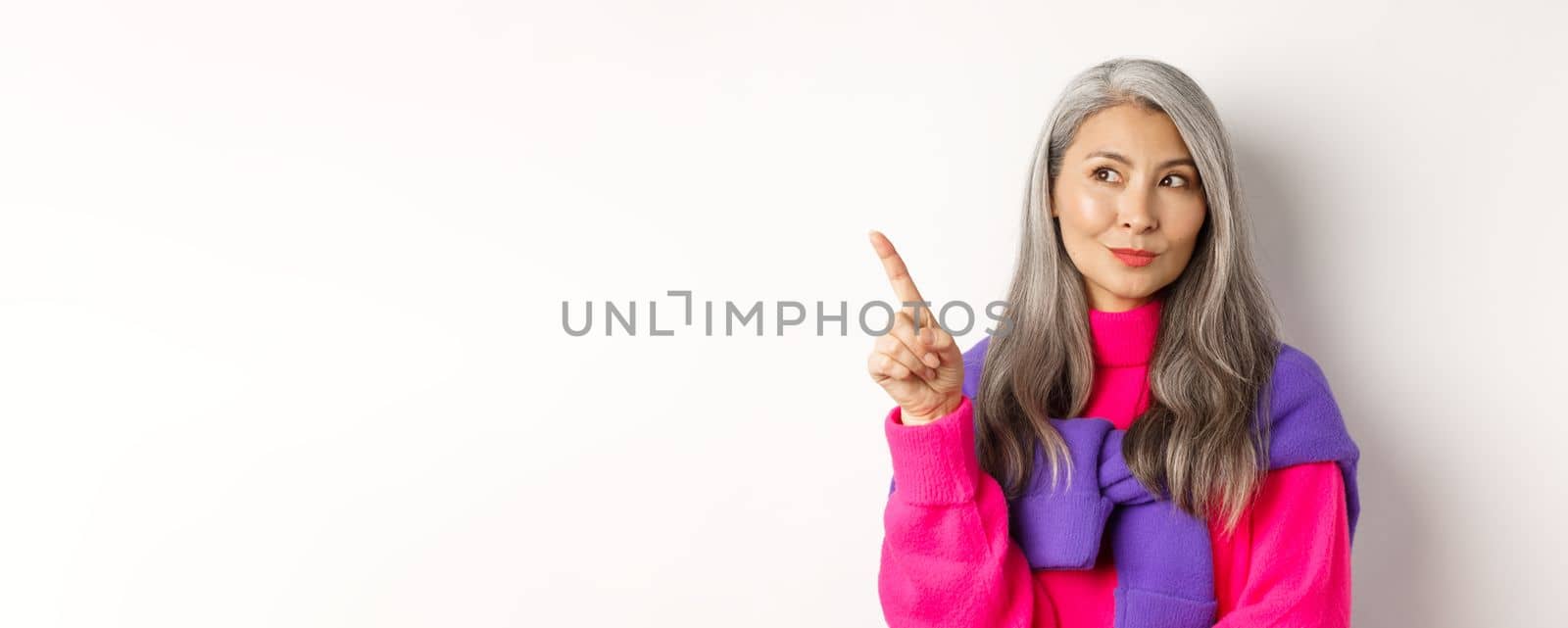 Close-up of stylish korean senior woman with grey hair looking and pointing upper left corner, showing promotion offer, standing over white background.