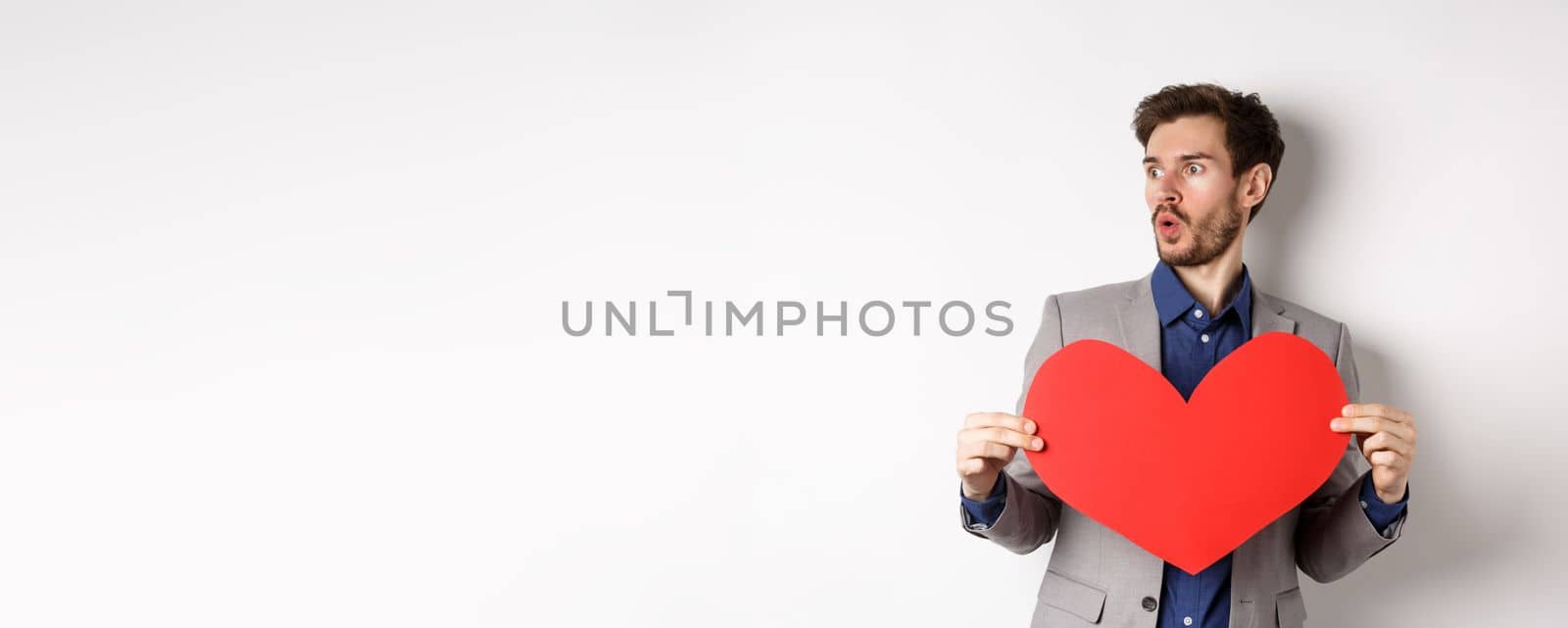 Man looking amazed at someone beautiful, holding big red heart cutout, standing on Valentines day over white background. Copy space