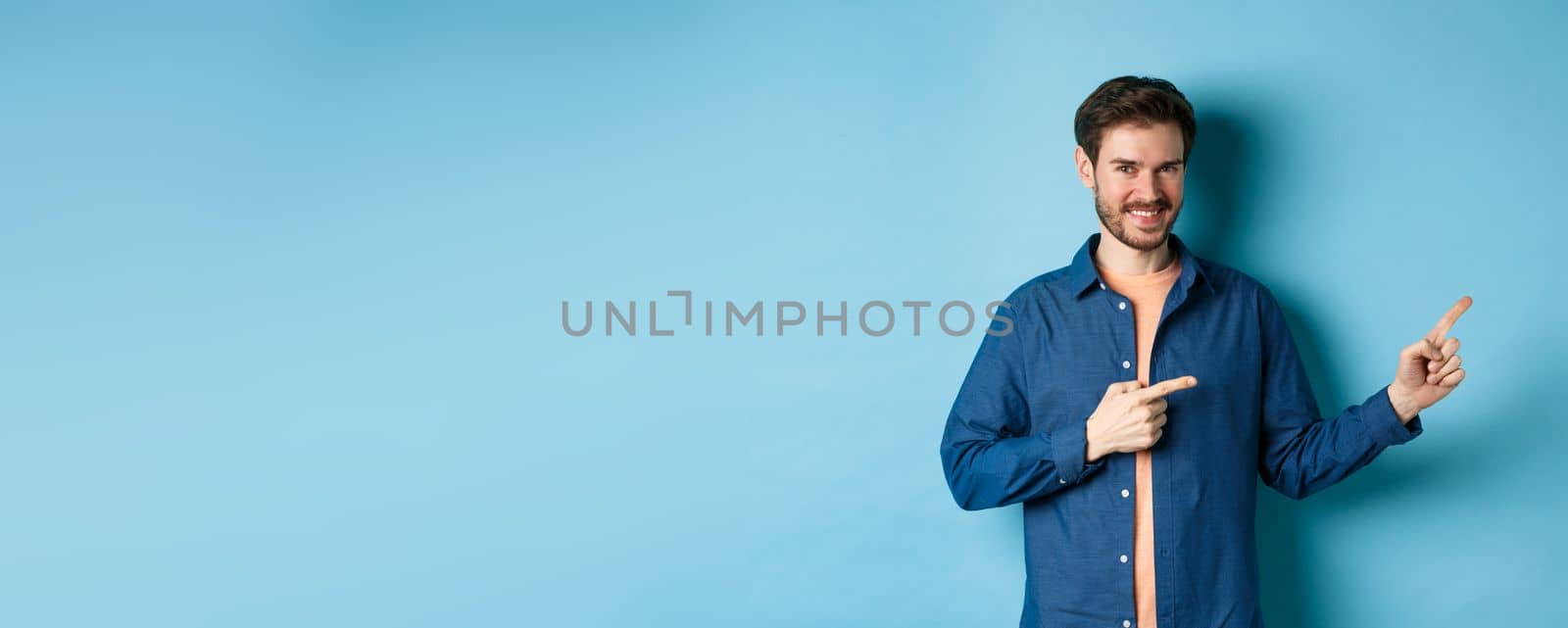 Smiling confident man showing logo, pointing fingers right at empty space and looking at camera, standing on blue background by Benzoix
