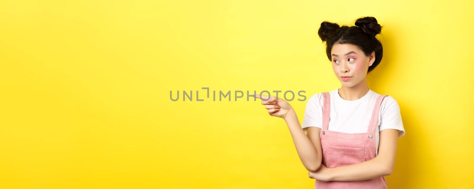 Portrait of stylish asian woman with makeup and summer clothes, pointing and looking left at logo, standing intrigued on yellow background by Benzoix