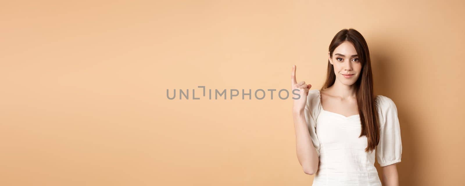 Attractive young woman show finger number one, smiling and looking confident, standing on beige background by Benzoix