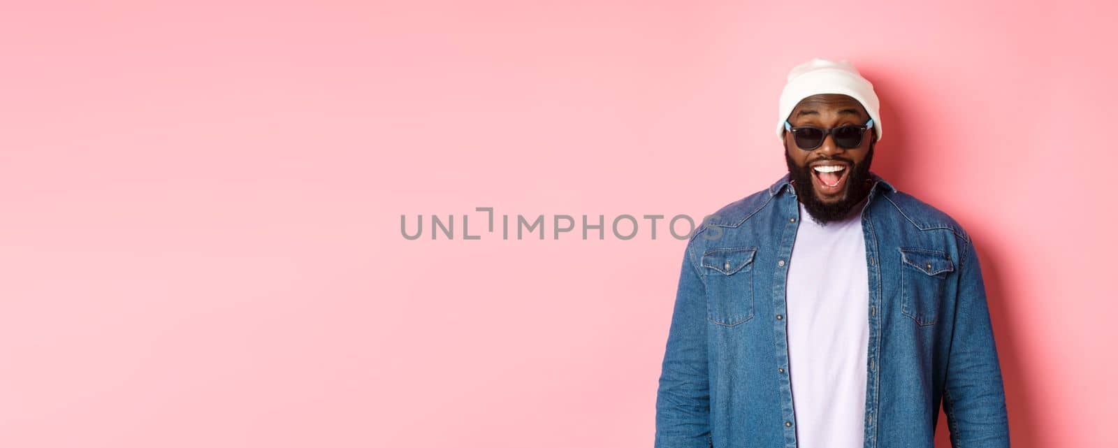Surprised and happy african-american hipster man in beanie and sunglasses, smiling and looking at camera, standing confident over pink background by Benzoix