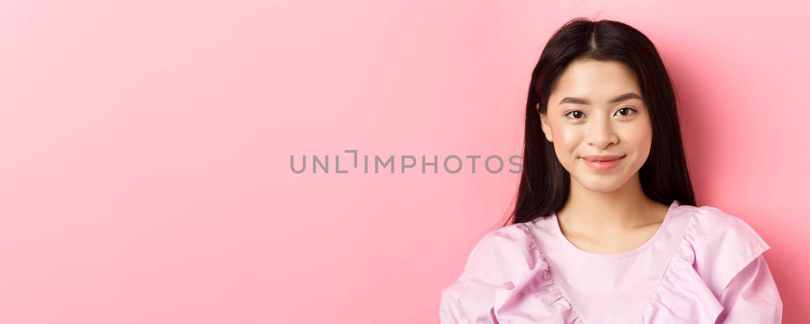 Close-up of beautiful asian woman looking happy, smiling cute at camera, standing in dress against pink background by Benzoix