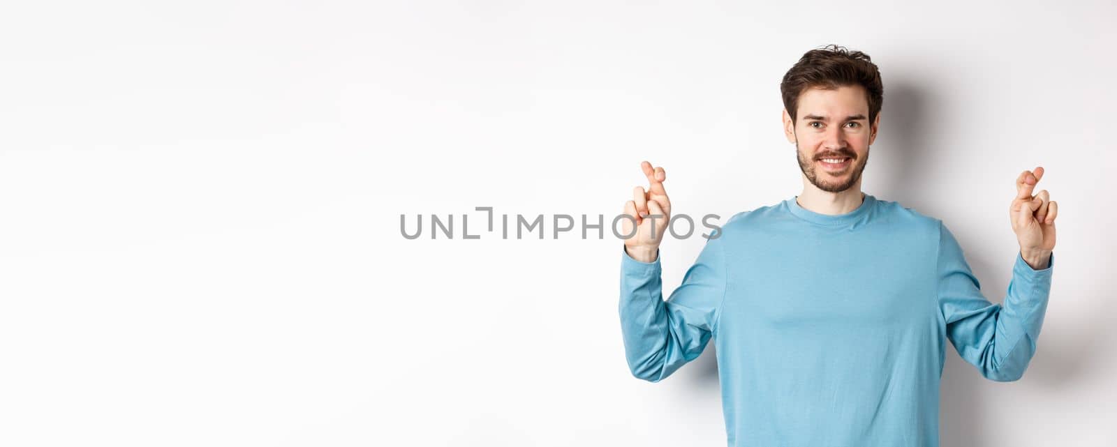 Optimistic smiling man cross fingers for good luck, making wish and waiting for results, standing over white background.