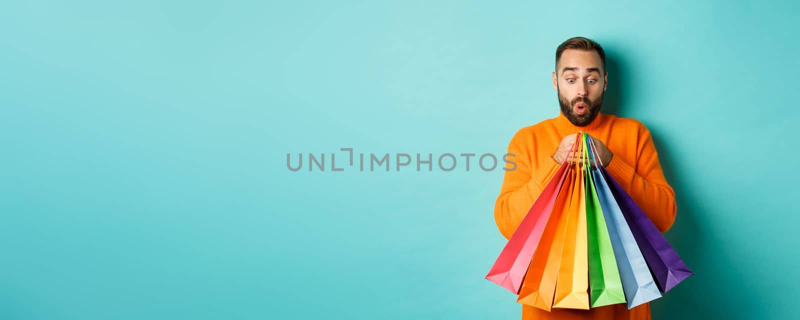 Excited man looking at shopping bags with purchases, standing amused against turquoise background by Benzoix