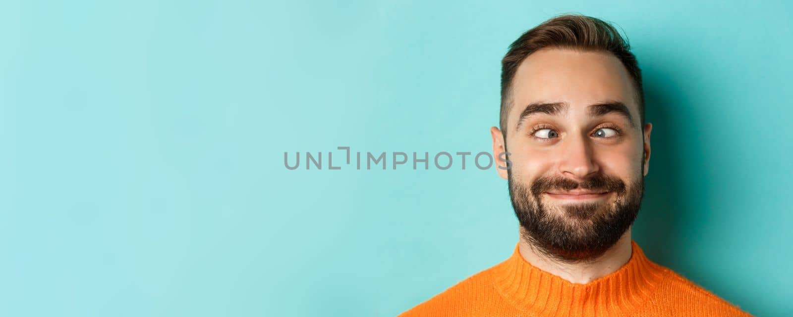 Headshot of funny caucasian man making faces, squinting and smiling silly, standing over light blue background.