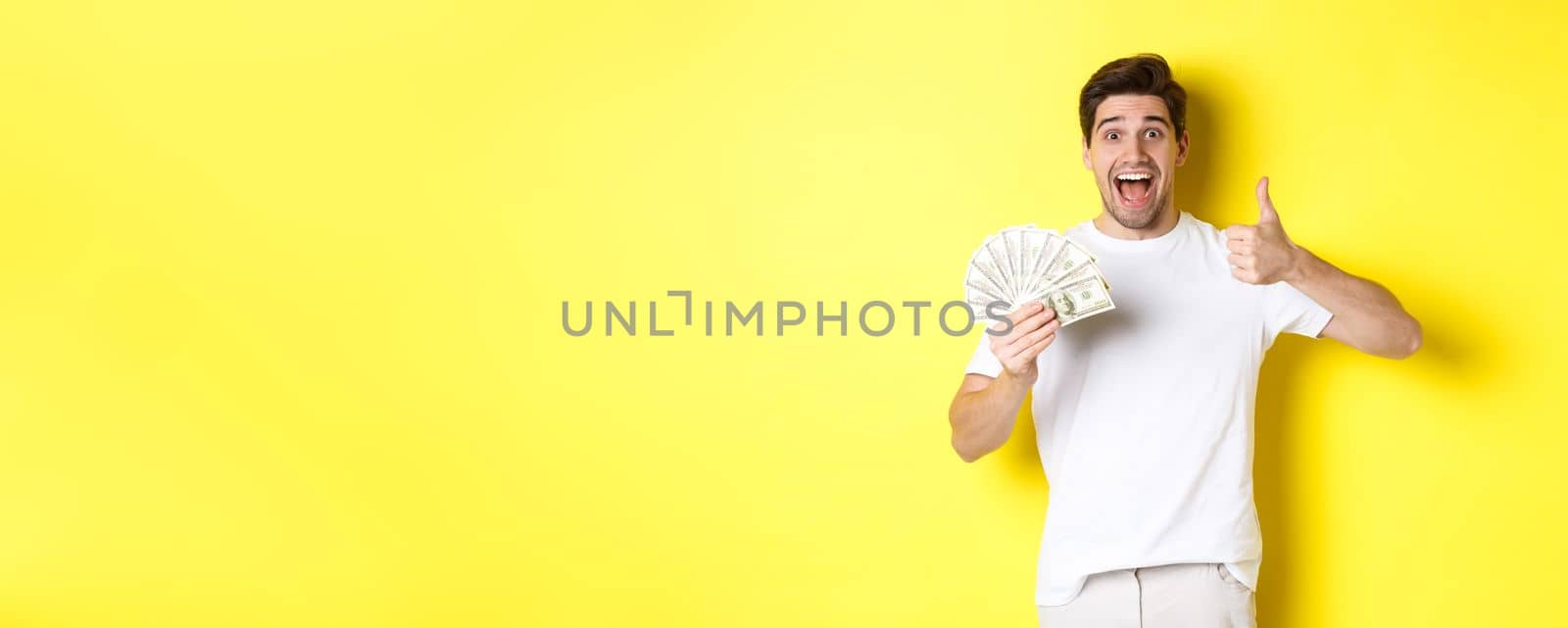 Excited man holding money, showing thumb-up in approval, got credit or loan, standing over yellow background.