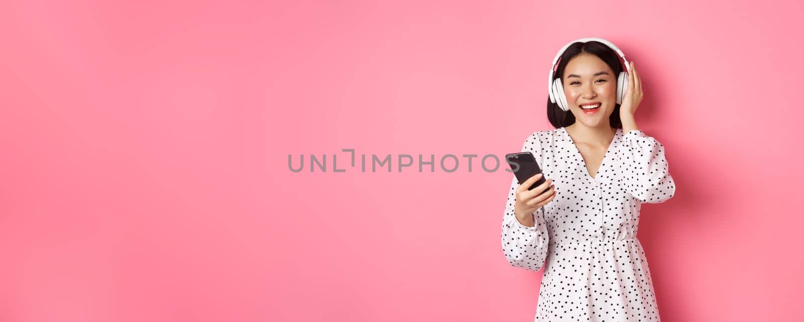 Beautiful asian woman listening music in headphones, using mobile phone, smiling happy at camera, standing over pink background by Benzoix