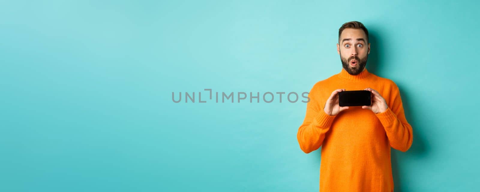 Impressed man showing smartphone screen, stare at camera amazed, demonstrate display, standing over light blue background by Benzoix