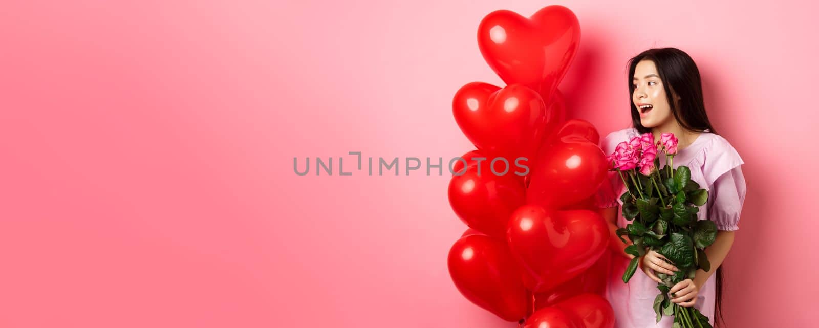 Portrait of asian teenage girl in love, holding flowers and looking at valentines day heart balloons, being on romantic date, pink background.