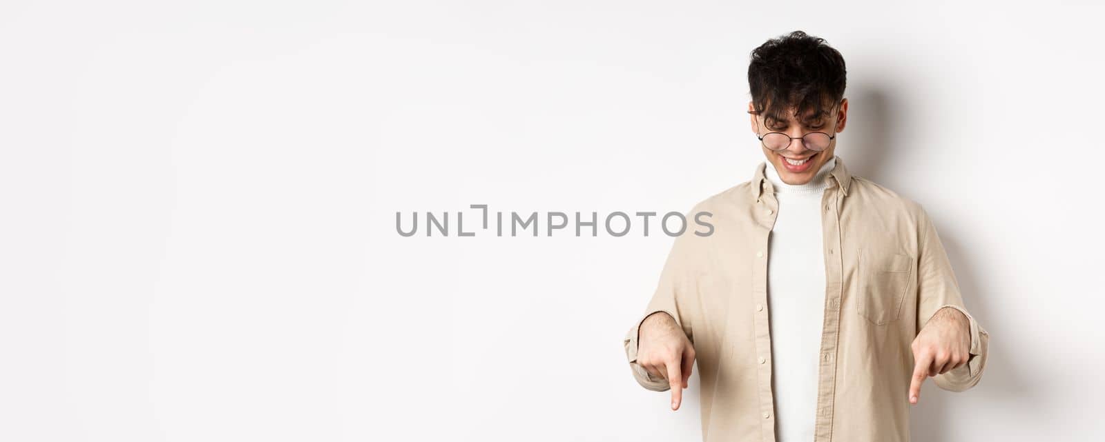 Stylish modern guy in glasses pointing, looking down with happy smile, showing banner, standing on white background.