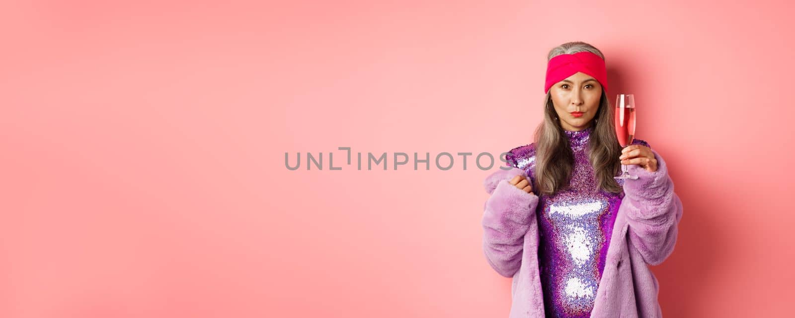 Fashionable asian senior female model raising glass of champagne, wearing trendy glitter dress and faux fur and looking at camera, congratulating with holiday, pink background.