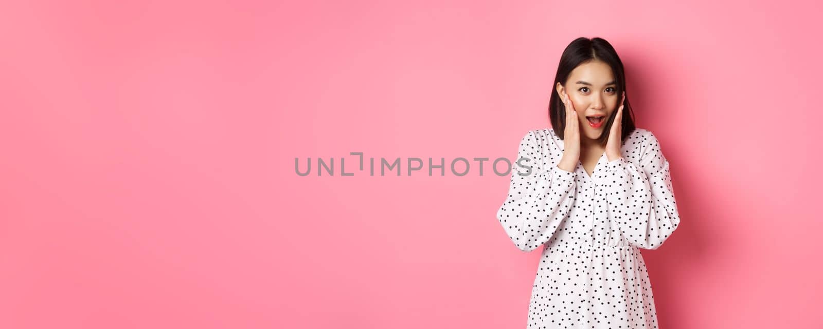 Young asian woman looking intrigued, gossiping and saying wow with curious face, looking interested at camera, standing over pink background by Benzoix