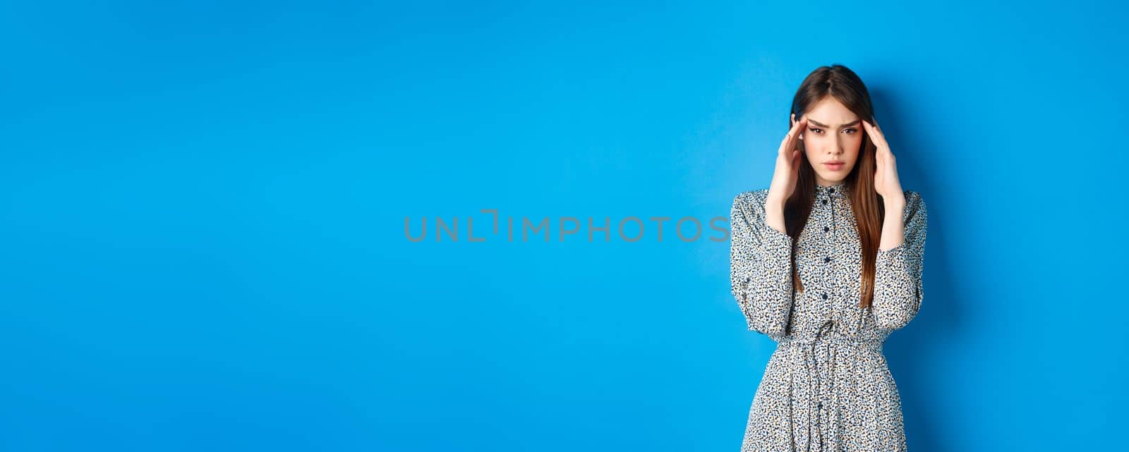 Woman trying to concentrate, touching head and squinting at camera, thinking or remember something, standing in dress on blue background by Benzoix