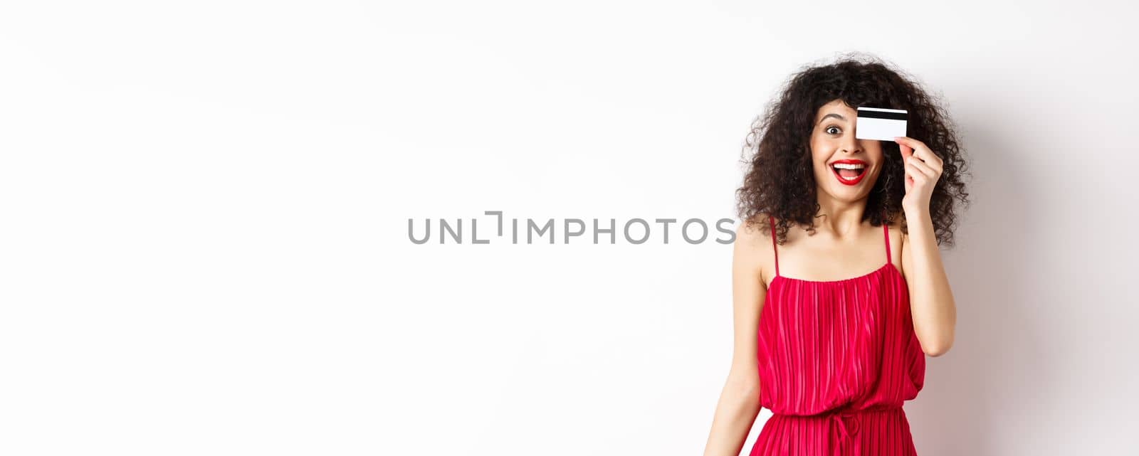 Shopping. Excited curly girl in red dress, showing plastic credit card and scream from joy, amazed with discounts and prices, standing against white background by Benzoix