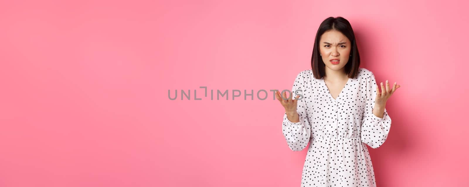 Angry and confused asian woman pointing hands at camera and grimacing furious, standing annoyed against pink background by Benzoix