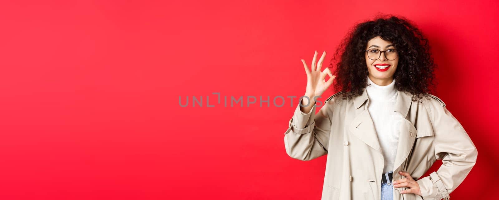 Portrait of confident trendy woman in glasses and trench coat, showing okay gesture to approve or agree with you, say yes, standing on red background.