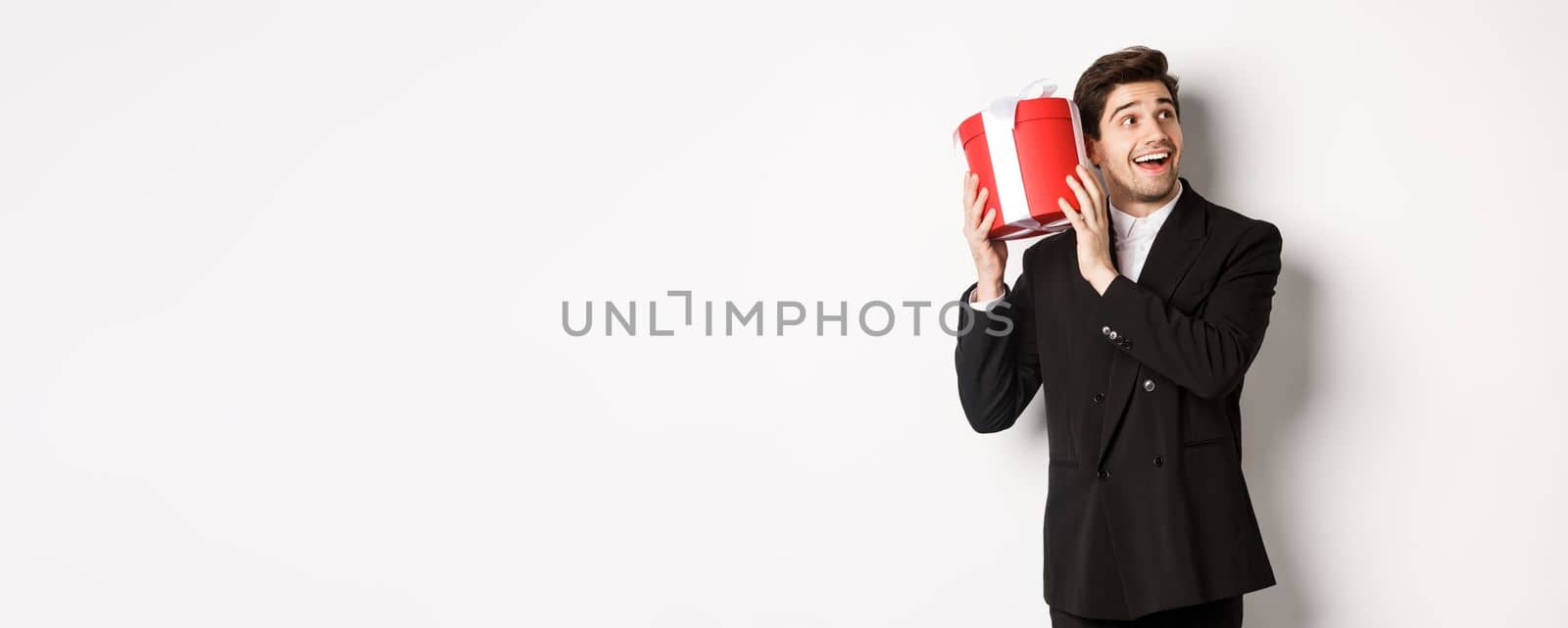 Concept of christmas holidays, celebration and lifestyle. Image of attractive man in black suit, shaking present to guess what inside, enjoying new year, standing against white background.
