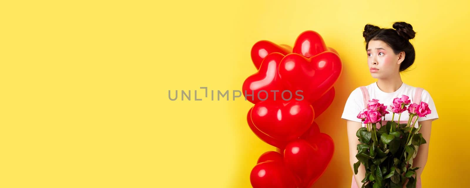 Happy Valentines day. Sad and lonely girl looking left upset, holding bouquet of roses, standing alone near red hearts balloons and frowning, yellow background.