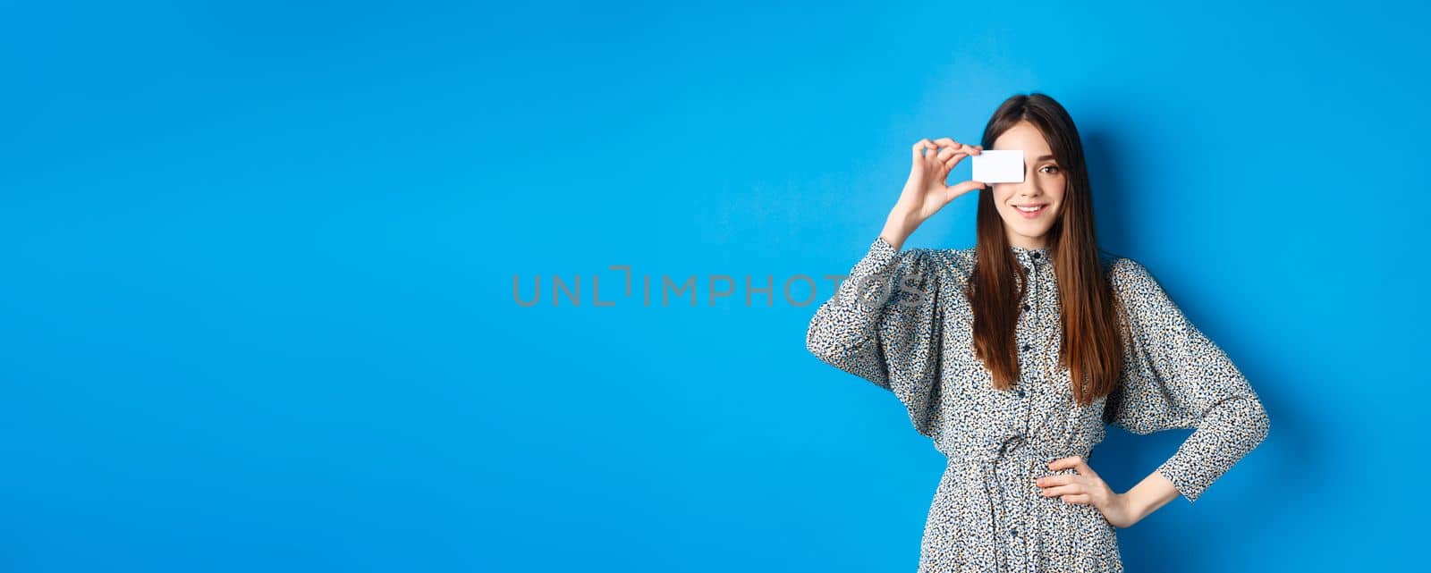 Shopping. Beautiful adult woman with natural long hair showing plastic credit card over eye, standing against blue background.