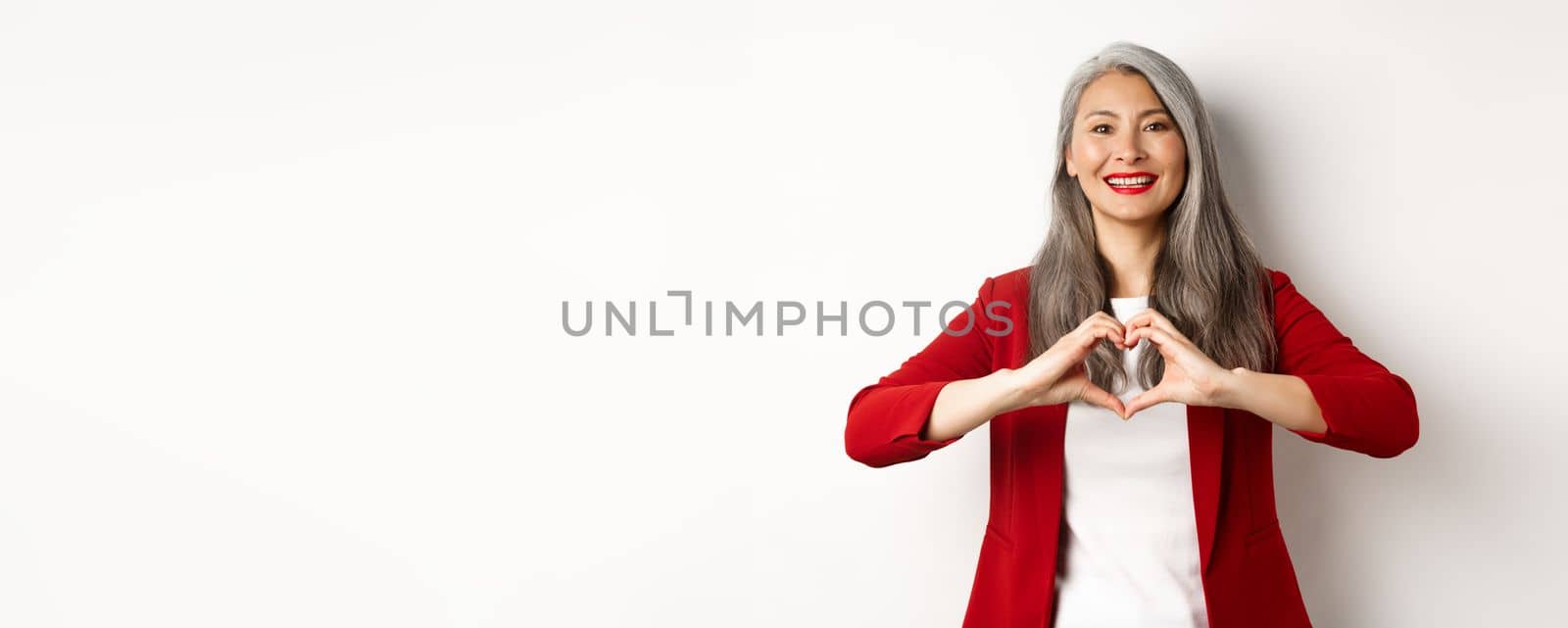 Beautiful asian mature woman in red blazer and makeup, showing heart sign and smiling, I love you gesture, standing over white background by Benzoix