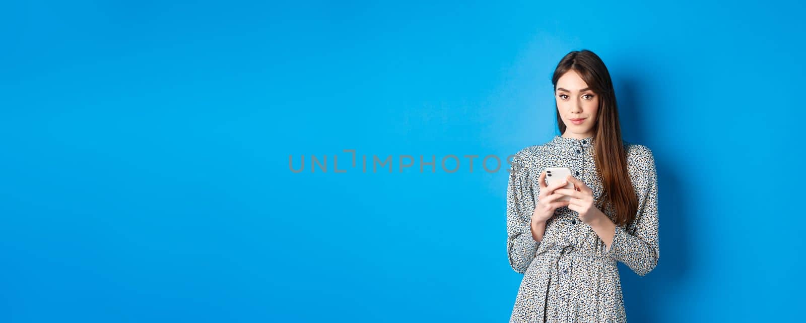 Stylish modern woman in dress, chatting on smartphone and smiling at camera, standing on blue background.