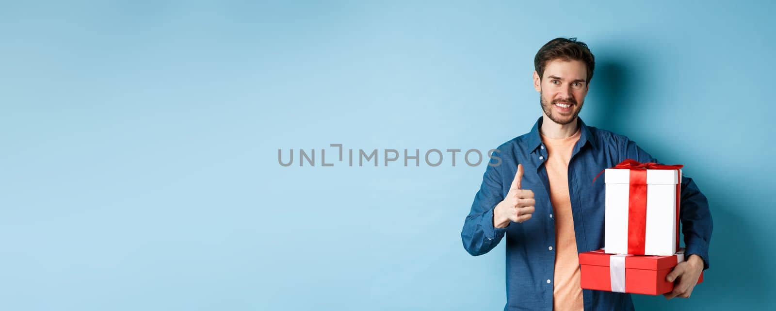 Smiling man holding romantic gifts and showing thumbs-up, celebrating Valentines day, buying presents for lover, standing over blue background.