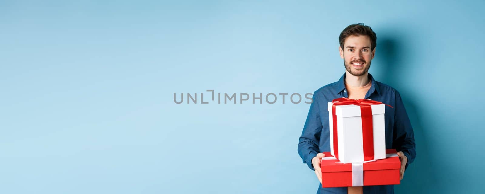 Happy valentines day. Handsome boyfriend holding gifts for lover on romantic date anniversary, standing over blue background.