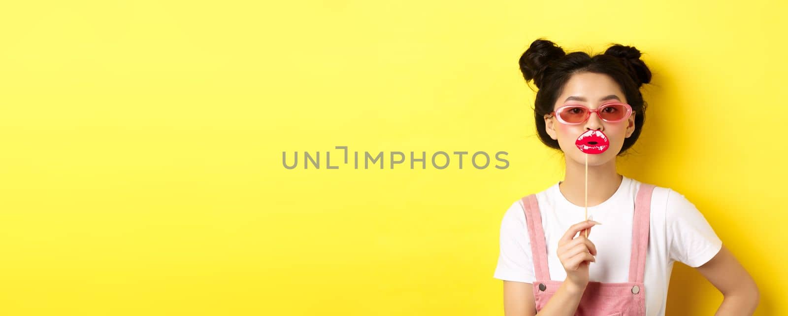 Summer and fashion concept. Stylish asian glam girl in sunglasses, holding party lips mask, standing on yellow background.