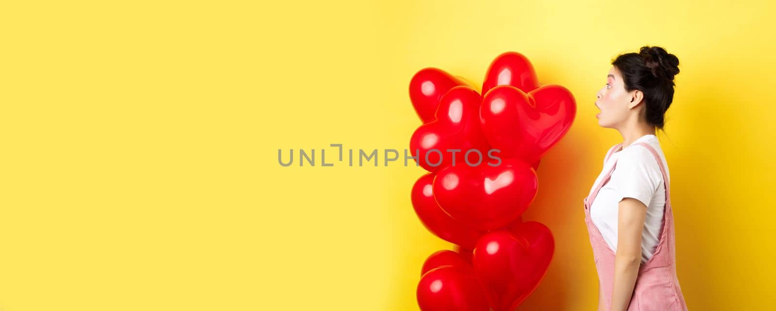 Valentines day and relationship concept. Profile of surprised asian girl staring left at logo, standing near heart balloons, yellow background by Benzoix