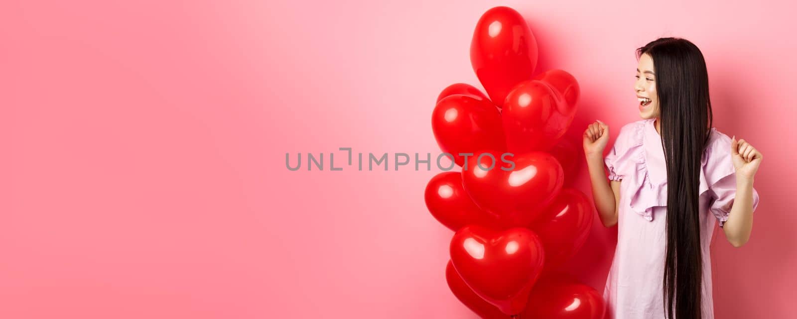 Asian teenage girl with long hair, cheering from valentines day romantic gift, looking at logo and smiling happy, jumping from joy near lovers gift heart balloons, pink background by Benzoix