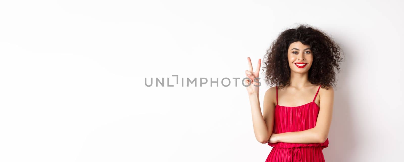 Attractive female model in red dress and makeup, showing number two and smiling, standing over white background by Benzoix
