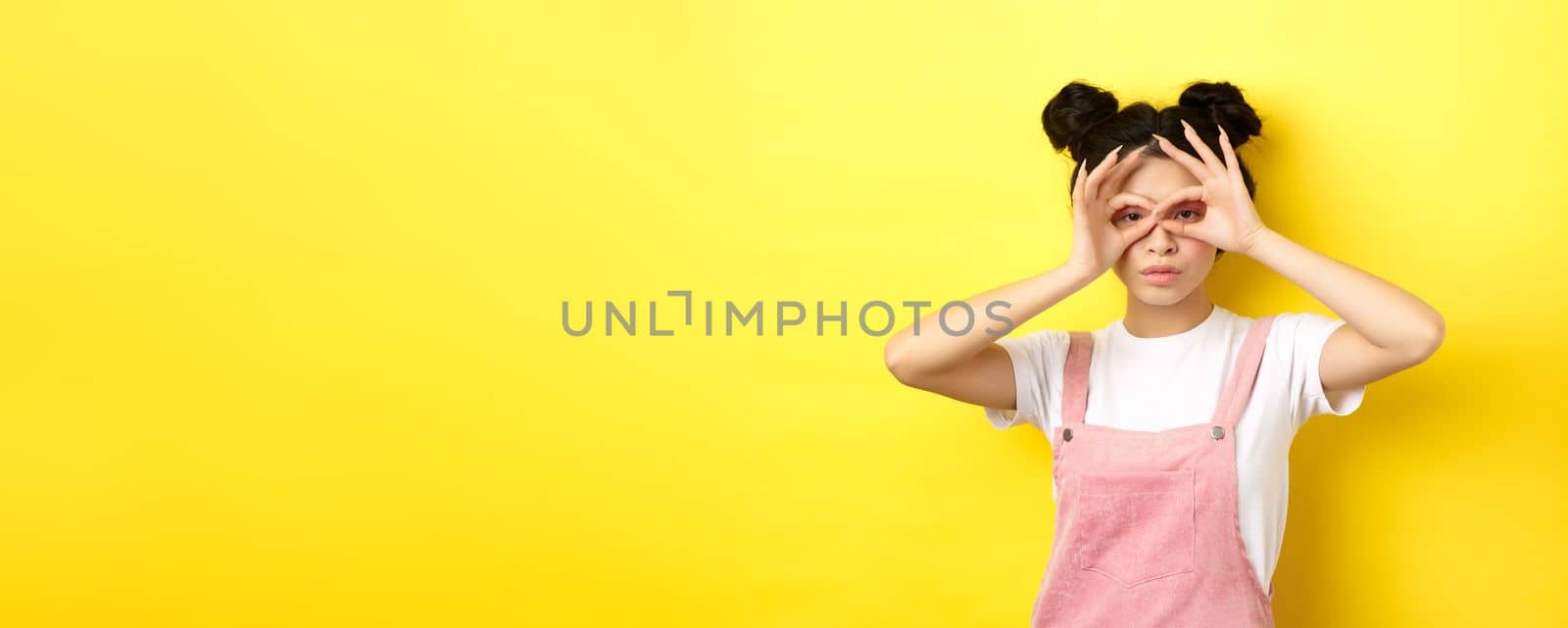 Funny asian girl look through hand binoculars, watching you, standing in summer clothes on yellow background.
