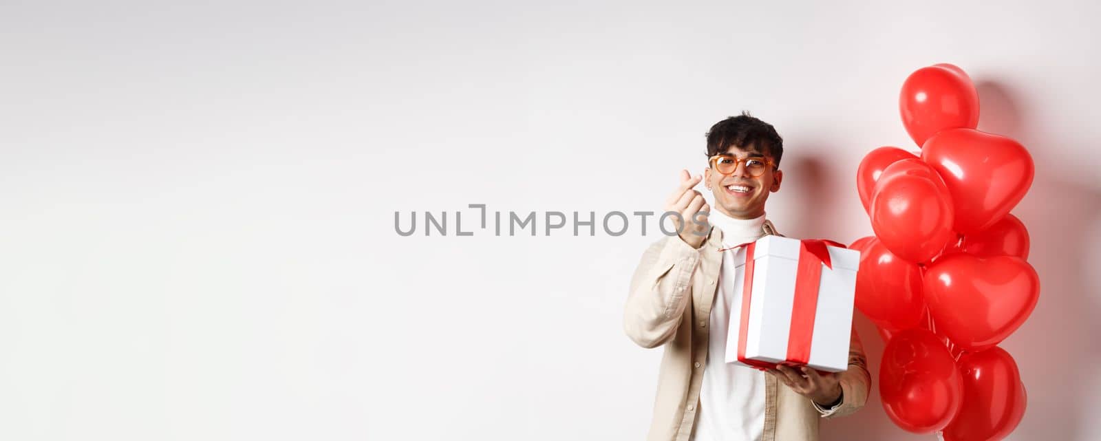 Valentines day and romance concept. Handsome young man showing surprise gift for lover and finger heart, smiling, standing near festive red balloons, white background by Benzoix