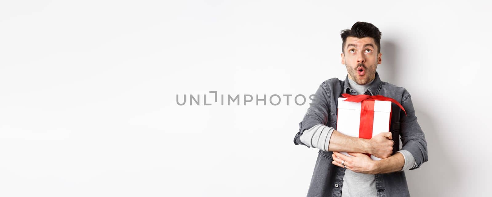 Surprised funny guy hugging big holiday gift and looking up, say wow amazed, checking out Valentines day special offer, standing on white background by Benzoix