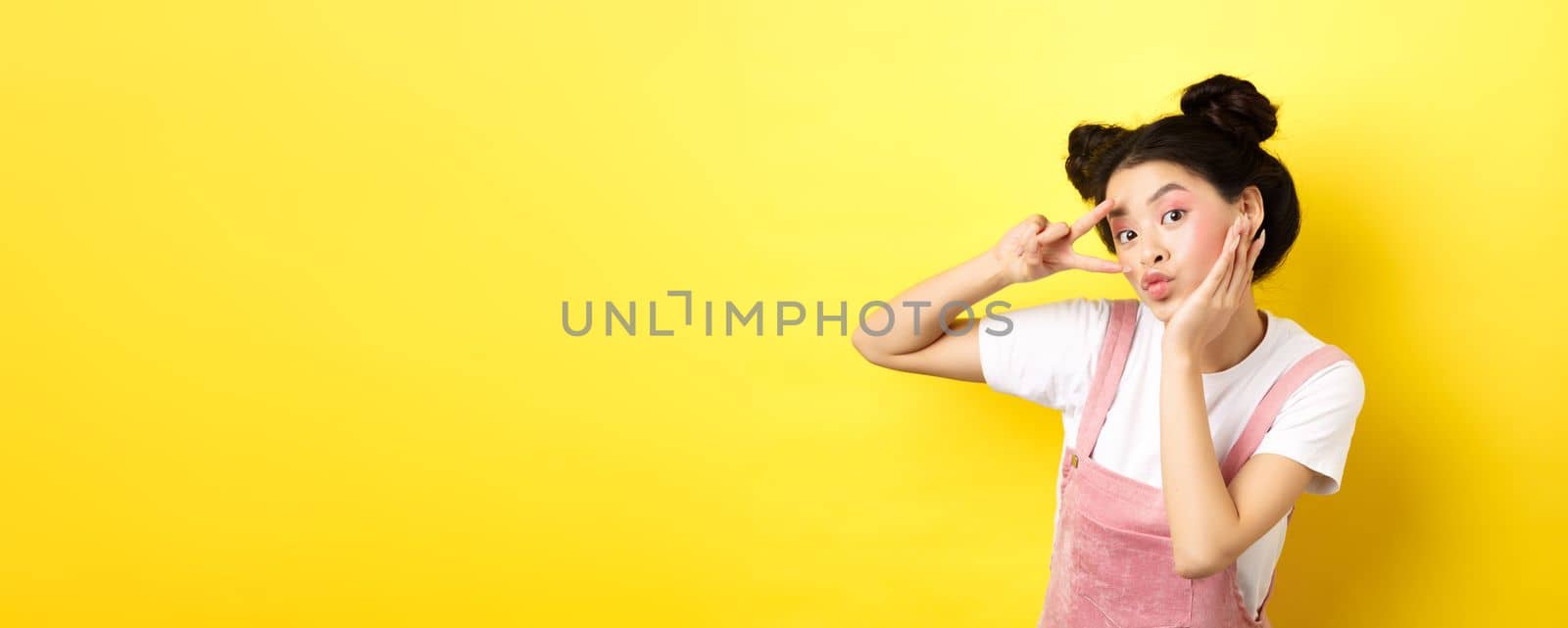 Beautiful asian girl showing v-sign and pouting cute, making silly face with makeup, standing on yellow background.