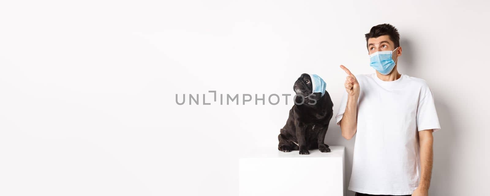 Covid-19, animals and quarantine concept. Young man and black dog wearing medical masks, pug and owner looking at upper left corner, white background by Benzoix
