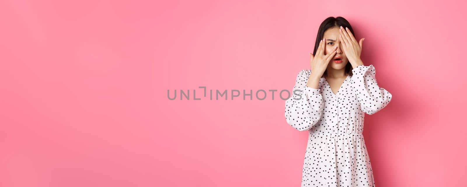 Disappointed asian woman peeking through fingers, open eyes and frowning displeased, staring with disdain at camera, standing over pink background.