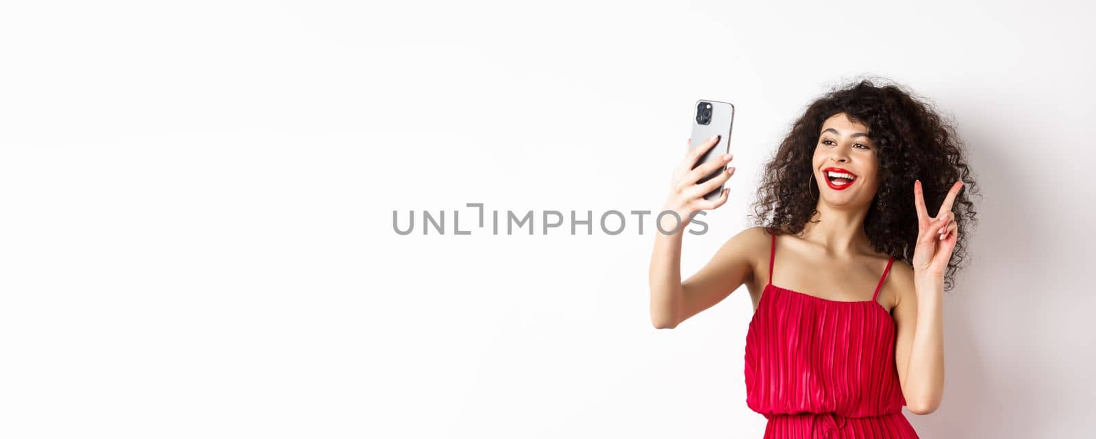 Elegant young woman in red dress taking selfie on smartphone, posing on event party, standing with mobile phone on white background by Benzoix