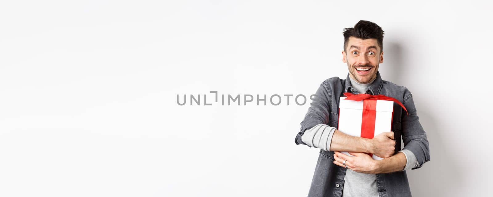 Happy caucasian man holding surprise gift, hugging present and smiling thankful, looking at camera with love and appreciation, celebrate valentines holiday, white background by Benzoix