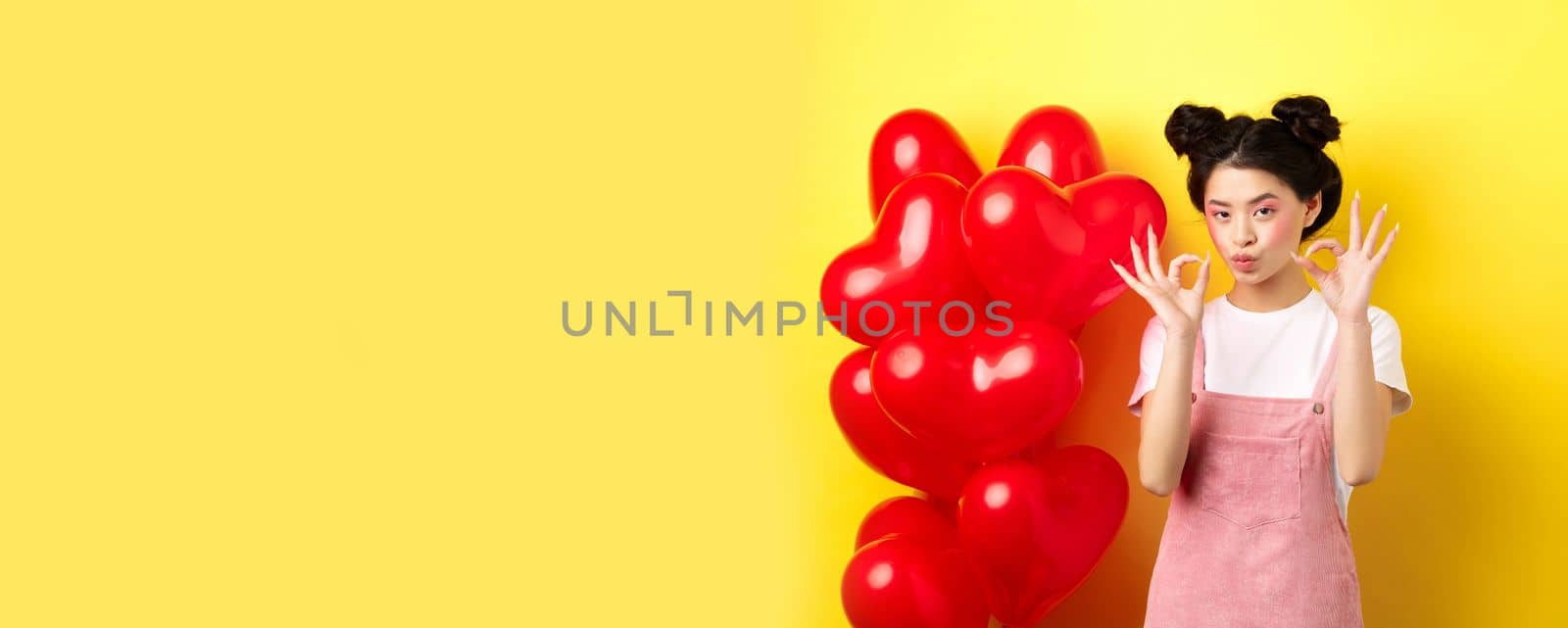 Cute young asian woman in stylish clothes and makeup, showing okay signs near st Valentines day heart balloons, praise good offer, standing on yellow background.